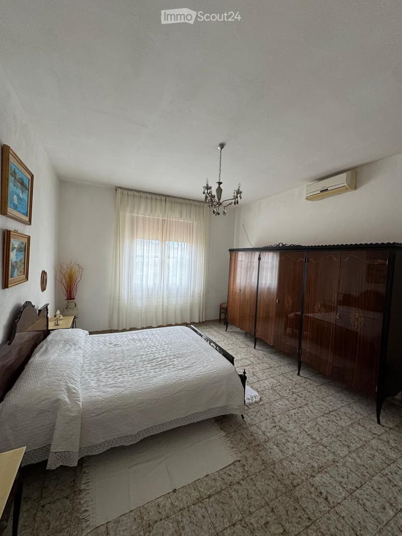 A well-lit bedroom with a large bed, a wooden wardrobe, a nightstand with a lamp, framed pictures on the wall, and a chandelier hanging from the ceiling.
