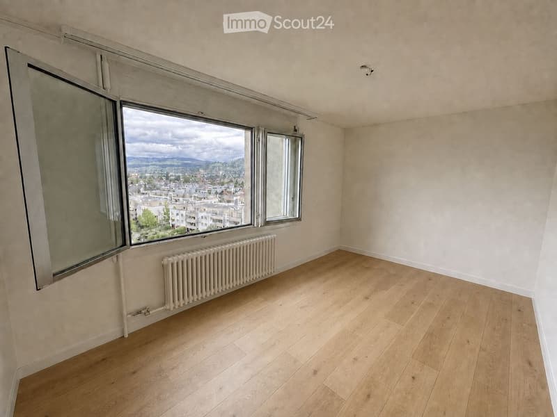 Empty room with light wooden flooring, large windows offering a cityscape view, and a white radiator under the windows.