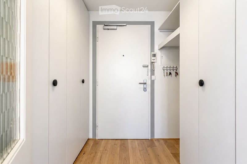 A modern, minimalist hallway with light wooden flooring, white walls, and a closed door leading to another room. A small built-in shelf unit with hooks for keys is visible on the right side.