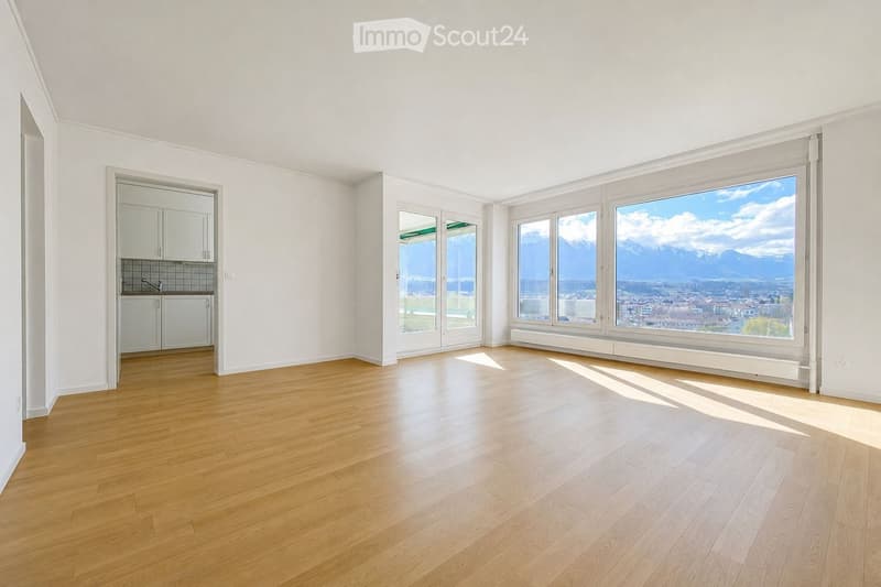 Empty room with wooden flooring, white walls, large windows offering a scenic mountain view, and a small kitchen area with cabinets and a sink.