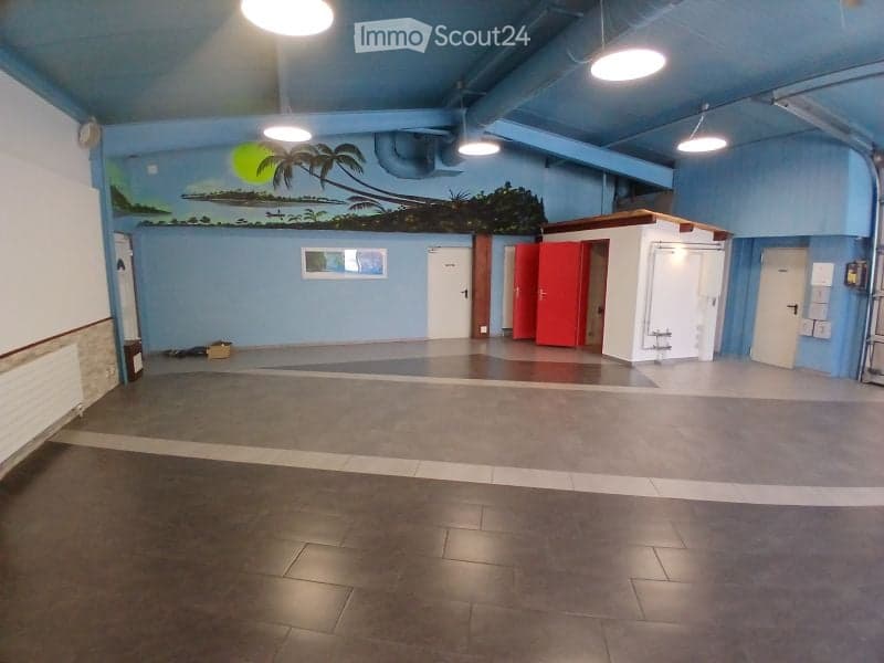 Empty room with light blue walls, a mural of a tropical scene, and several doors including red and white ones. The floor is tiled, and the ceiling has multiple hanging lights.