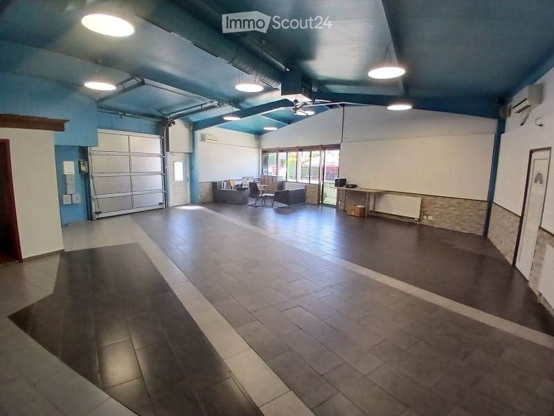 Large, open garage space with a polished concrete floor, blue-painted ceiling, and several overhead lights. The area includes a large roll-up door, a few storage items, and a small seating area with chairs and a table near the back.