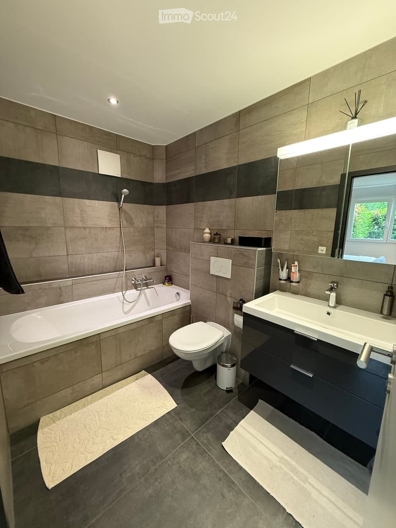 Modern bathroom with a bathtub, toilet, sink, and large mirror. Features tiled walls, a shower head, and a small shelf with toiletries.