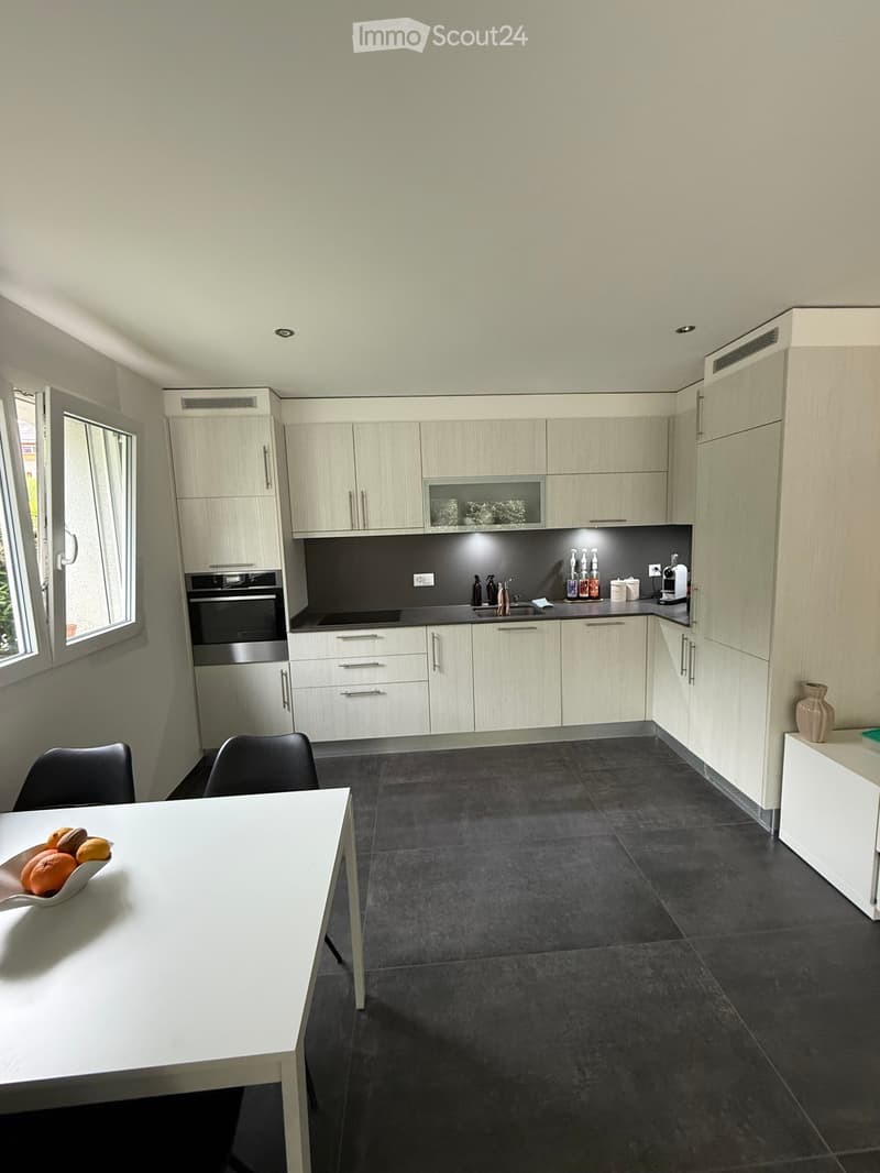 Modern kitchen with light wood cabinets, black countertops, and stainless steel appliances. Features a white dining table with chairs, a bowl of fruit, and a vase on a sideboard.
