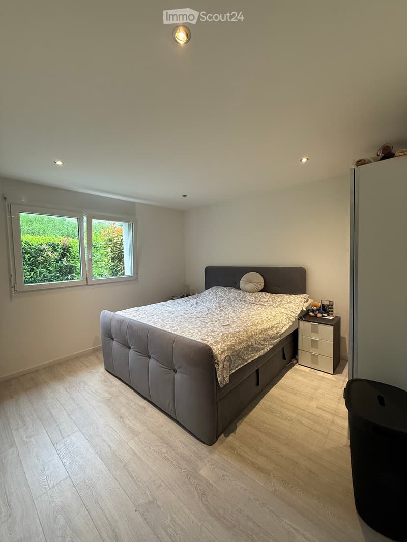 Modern bedroom with a large bed, nightstand, and a view of greenery through the window.