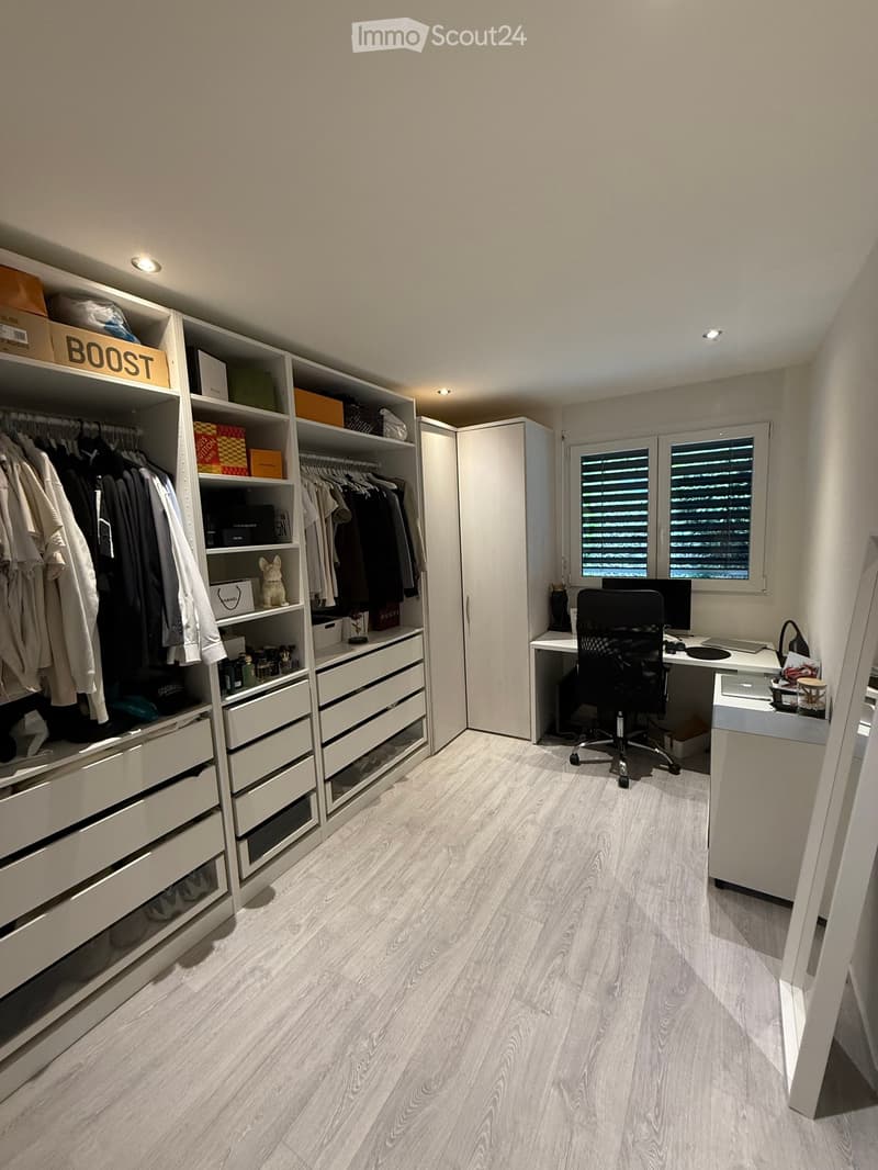 A modern, well-organized walk-in closet with built-in wardrobe, drawers, and shelves. A desk with a chair and laptop is positioned near the window, with storage boxes and decorative items visible.