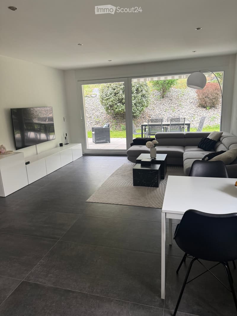 Modern living room with grey sectional sofa, coffee table, dining table, chairs, TV, and floor lamp. Large sliding glass doors lead to outdoor patio.