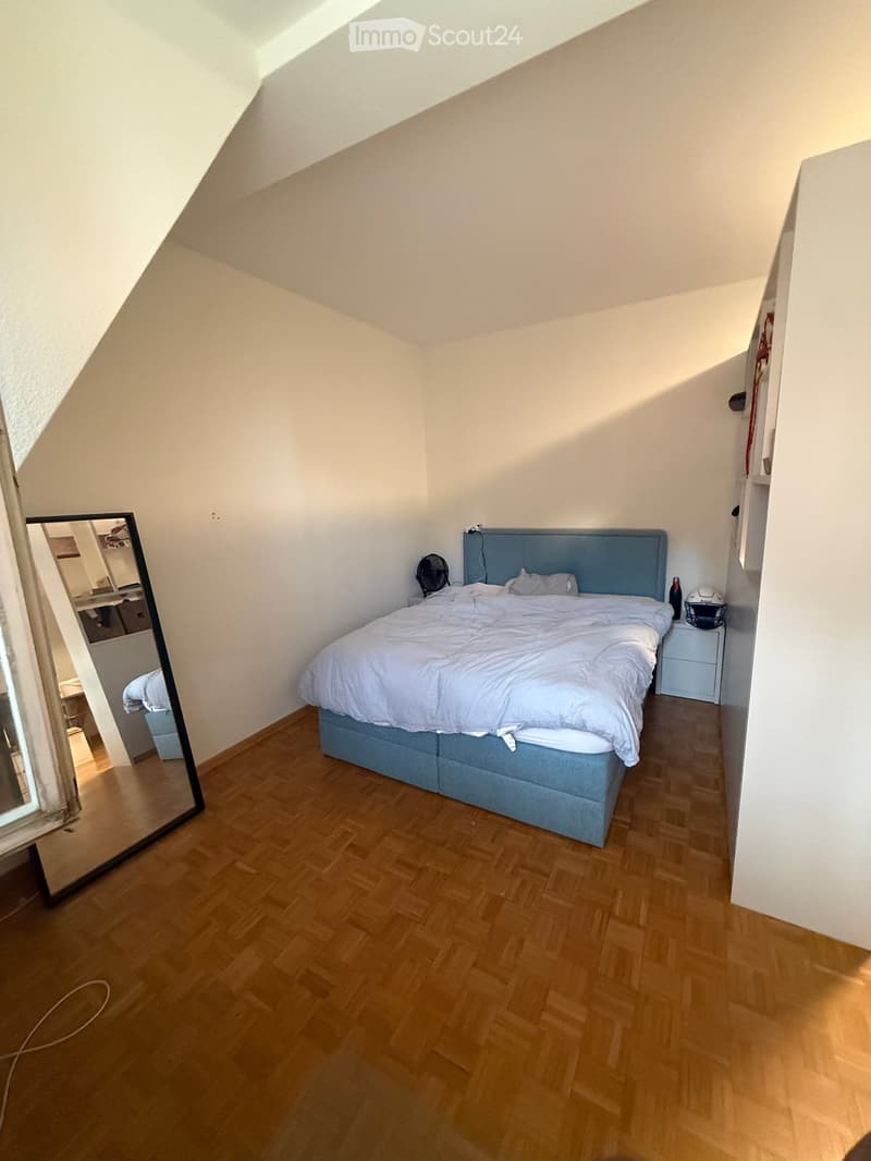 A small, well-lit bedroom with wooden flooring, a blue headboard bed, a nightstand, a fan, and a mirror on the wall.