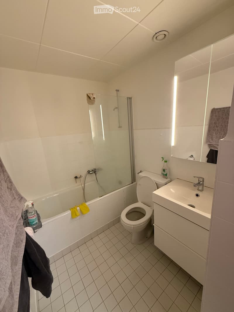 Modern bathroom with white tiles, bathtub, toilet, sink, and mirror. Includes towels and cleaning supplies.