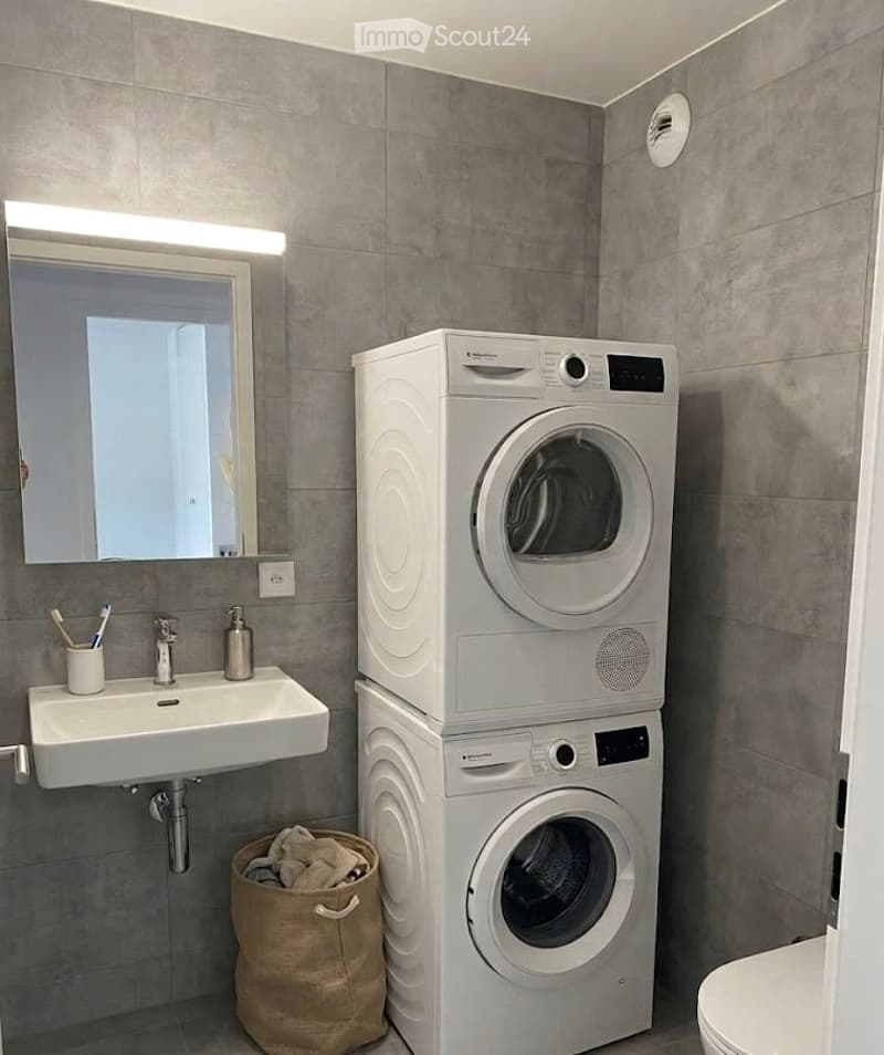 Modern bathroom with grey tiled walls, a white sink with a mirror, a washing machine and dryer stacked, a toilet, and a laundry bag on the floor.