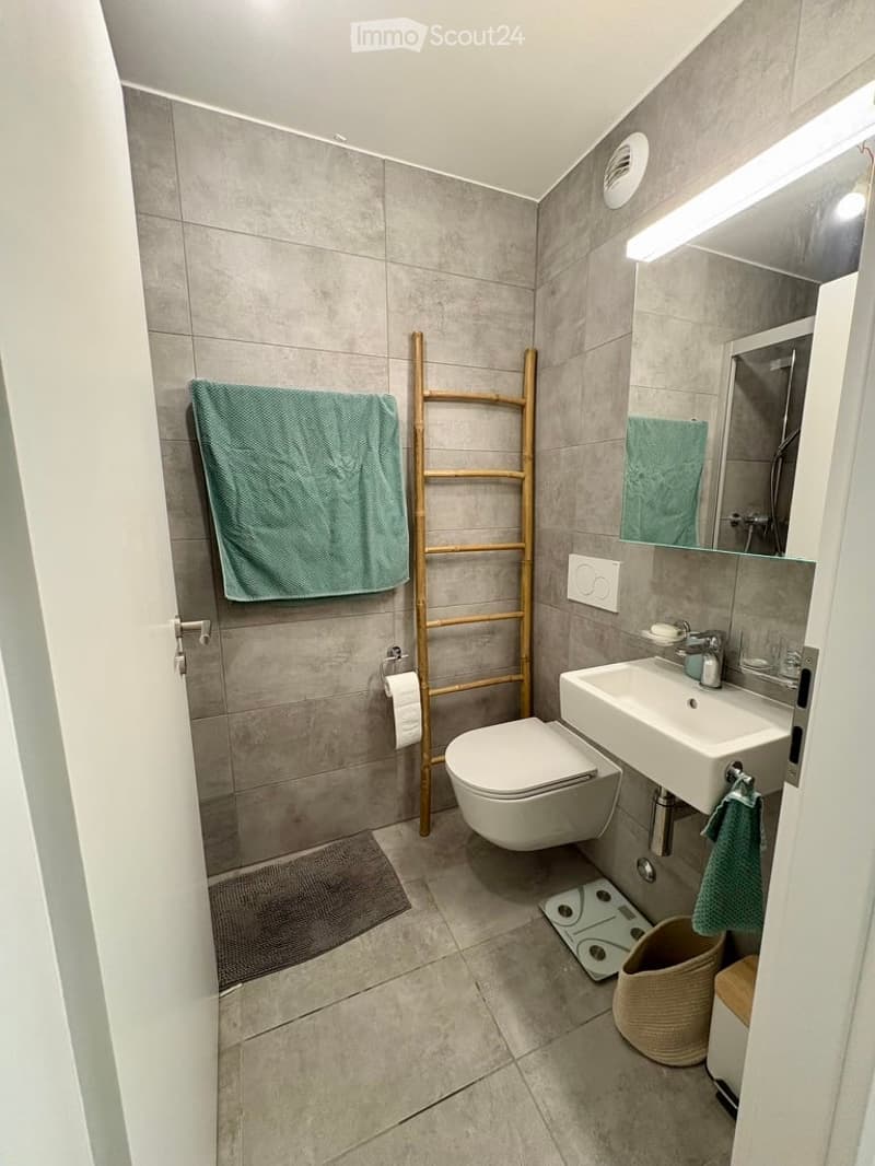 Modern bathroom with grey tiled walls and floor, white toilet, sink, and mirror, green towels, wooden ladder, and a woven basket on the floor.