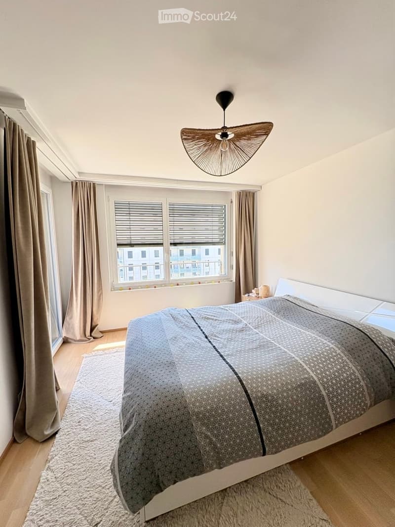 Modern bedroom with a large bed, geometric patterned bedspread, nightstand, and a stylish hanging lamp. The room features wooden flooring, large windows with blinds, and neutral-colored curtains.