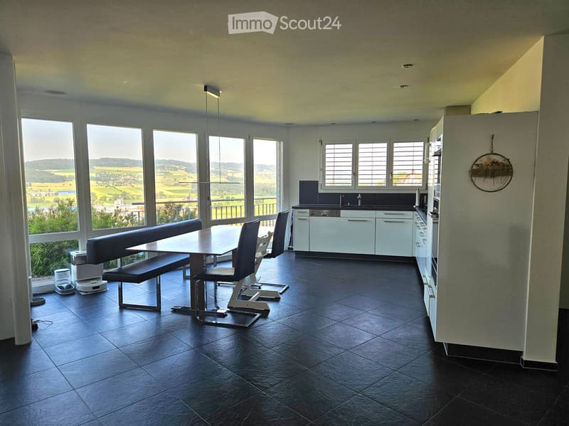 Modern kitchen and dining area with large windows offering a scenic view, featuring a dark tiled floor, a long dining table with bench seating, and sleek white kitchen cabinets.