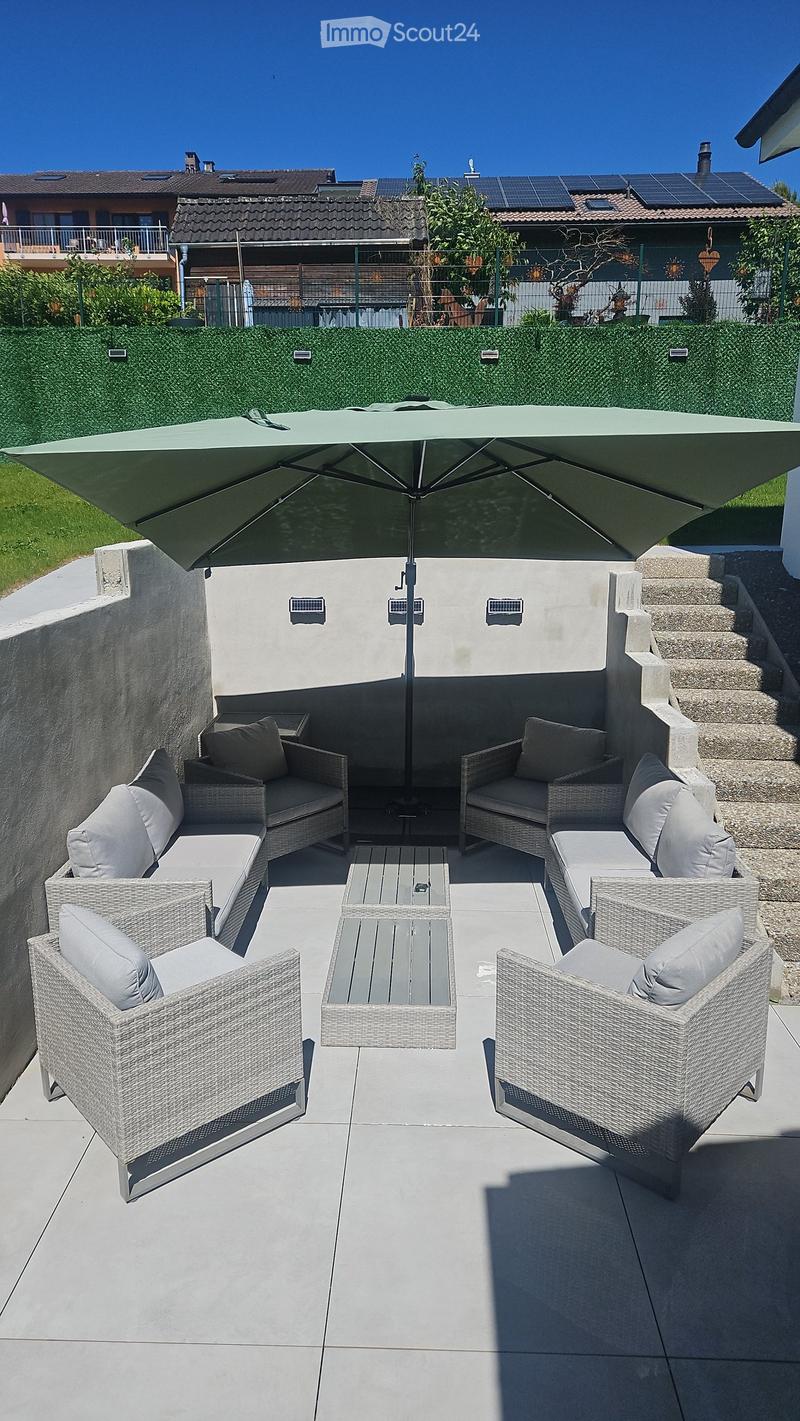 A modern outdoor patio with a large umbrella, wicker furniture including sofas and chairs, and a small coffee table.