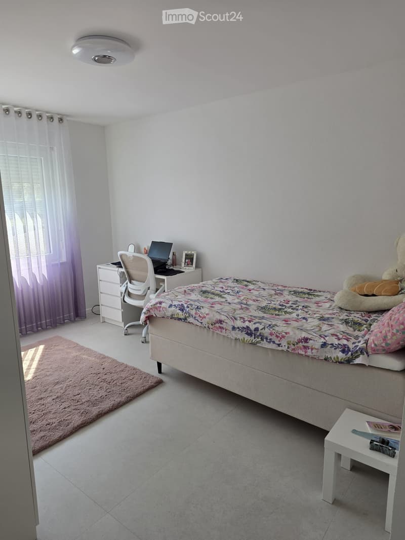 A well-lit bedroom with a bed, desk, chair, laptop, and a small white stool. The room features a floral bedspread, a teddy bear, and a picture frame on the desk.