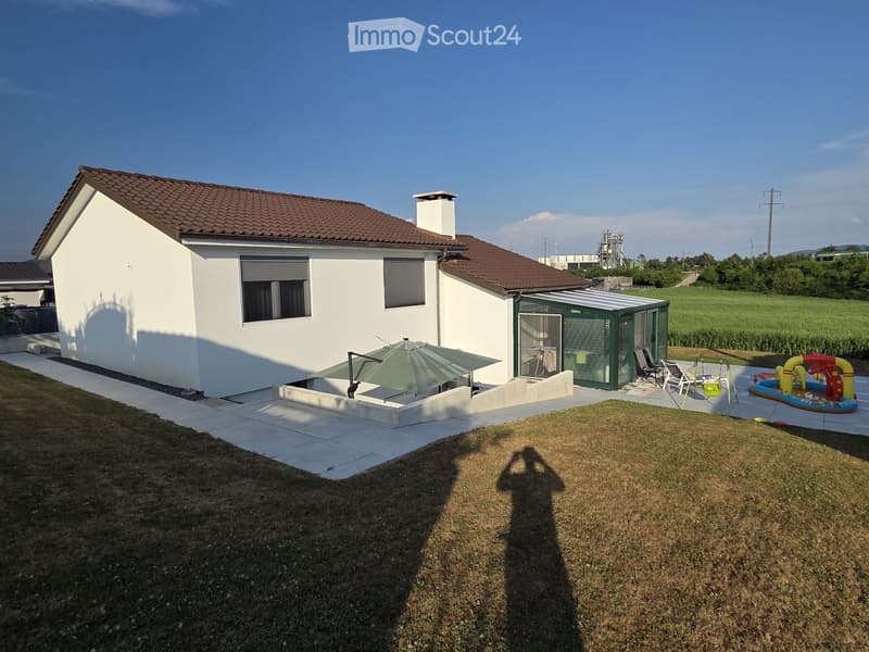Single-story white house with a tiled roof, surrounded by a grassy lawn and a patio area with outdoor furniture and a small inflatable pool.