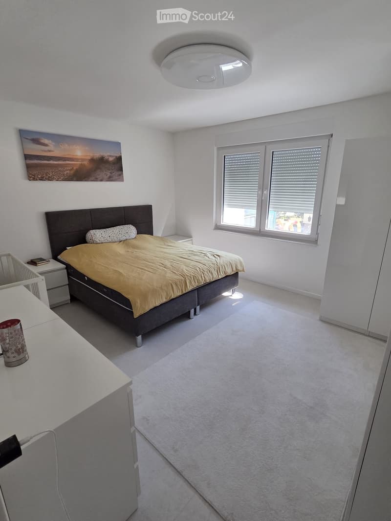 Modern bedroom with a large bed, nightstand, picture frame, and a cup on a white surface. The room features white walls, a large window with blinds, and a carpeted floor.