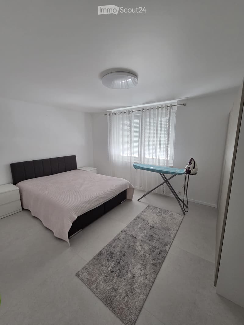 Bedroom with a bed, nightstand, ironing board, iron, and a rug. The room has white walls, a large window with curtains, and a modern ceiling light.