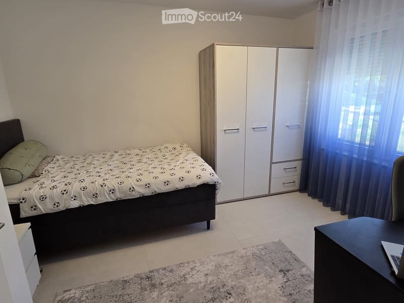 A modern bedroom with a bed, wardrobe, and a small table with a laptop. The room features light-colored walls, a tiled floor, and a large window with curtains.