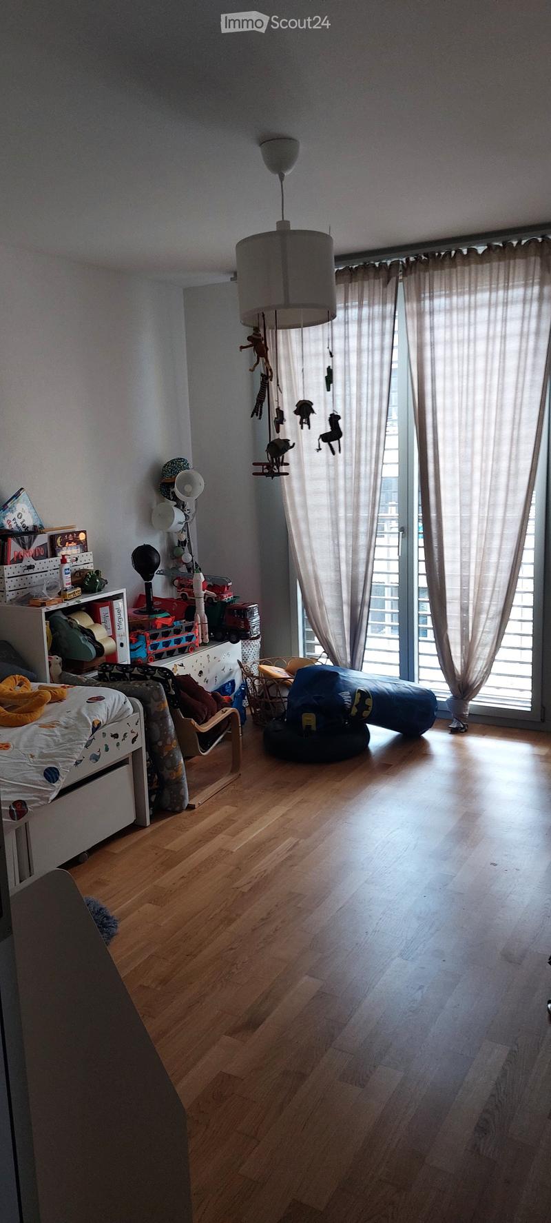 Brightly lit children's bedroom with wooden flooring, large window with curtains, bed with colorful bedding, shelves with toys and books, hanging toys, and a small table with a chair.