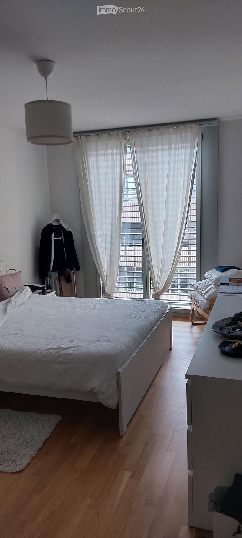 Bright bedroom with wooden flooring, large window with white curtains, bed with white bedding, nightstand, chair, and a hanging lamp.