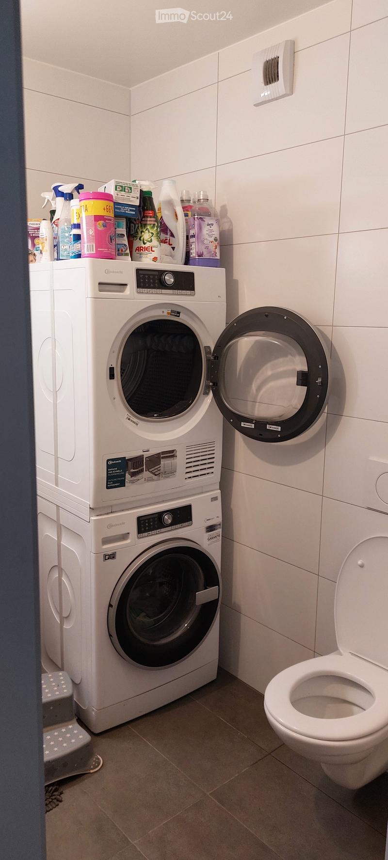 Bathroom with a washing machine, dryer, and toilet. Cleaning supplies are placed on top of the washing machine.