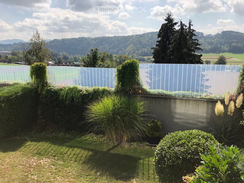Well-maintained garden with a variety of plants, shrubs, and a modern glass fence, overlooking a scenic valley with trees and hills in the background.