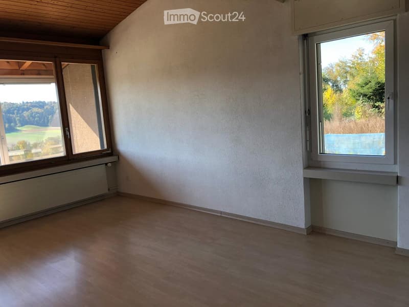 Empty room with wooden flooring, white walls, and large windows offering views of greenery and a body of water.