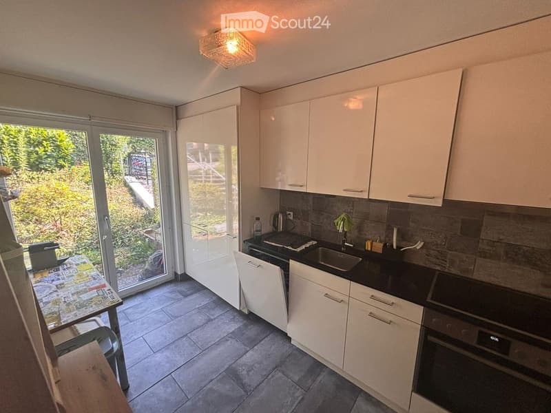 Modern kitchen with white cabinets, black countertops, and stainless steel appliances. Features a sink, oven, and a small table with a chair near the window.