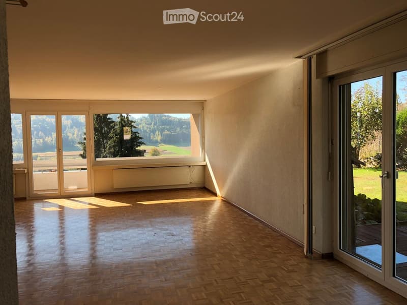 Empty room with wooden flooring, large windows, and sliding glass doors leading to a balcony and outdoor area.