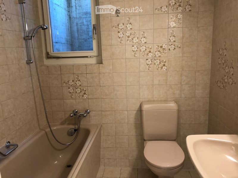 Bathroom with beige tiled walls, a bathtub with a handheld shower, a toilet, and a sink. The tiles feature a floral pattern.