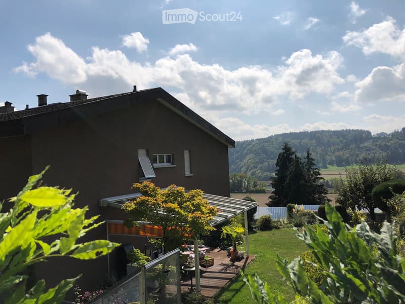 A house with a well-maintained garden, featuring a wooden deck, potted plants, and a scenic view of a hilly landscape with trees and fields.