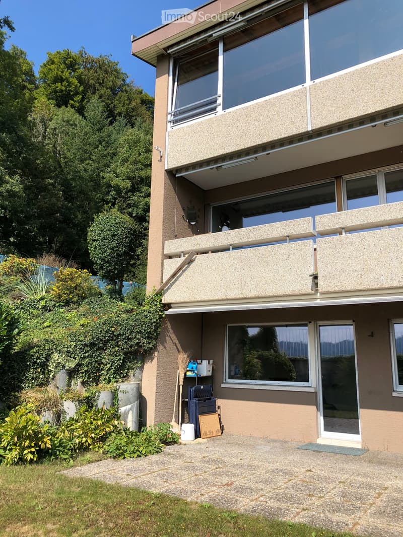 Two-story residential building with large windows, balconies, and a small garden area with plants and a concrete driveway.