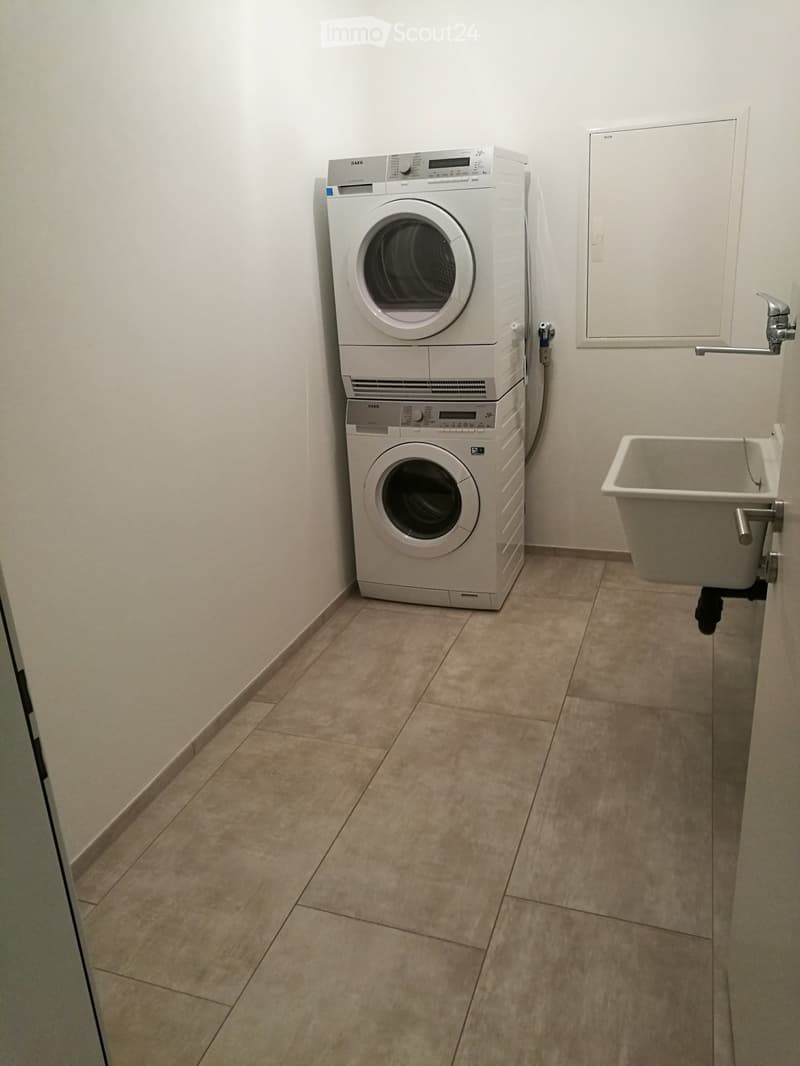 A small utility room with tiled flooring, white walls, and a stacked washing machine and dryer unit. A small sink is mounted on the wall to the right.