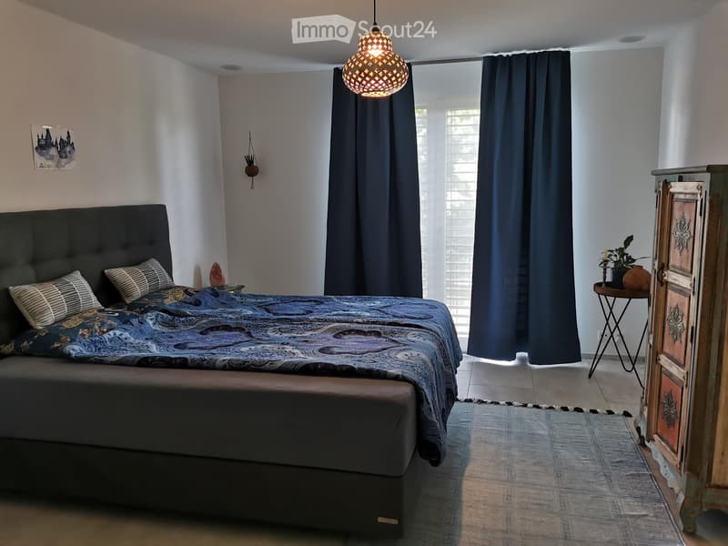 Modern bedroom with a large bed, patterned bedding, nightstand, pendant light, side table, and decorative plant.