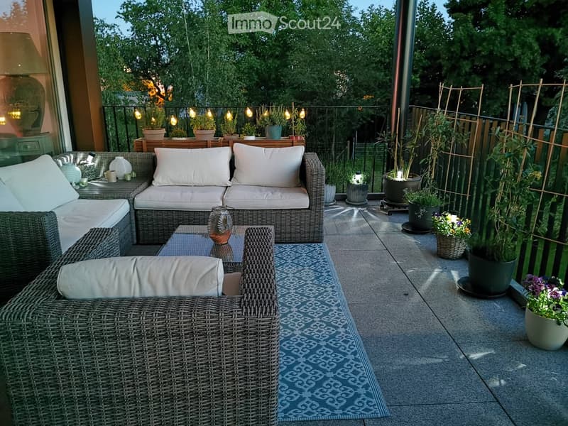 A well-furnished balcony with wicker furniture, a blue patterned rug, potted plants, and string lights.