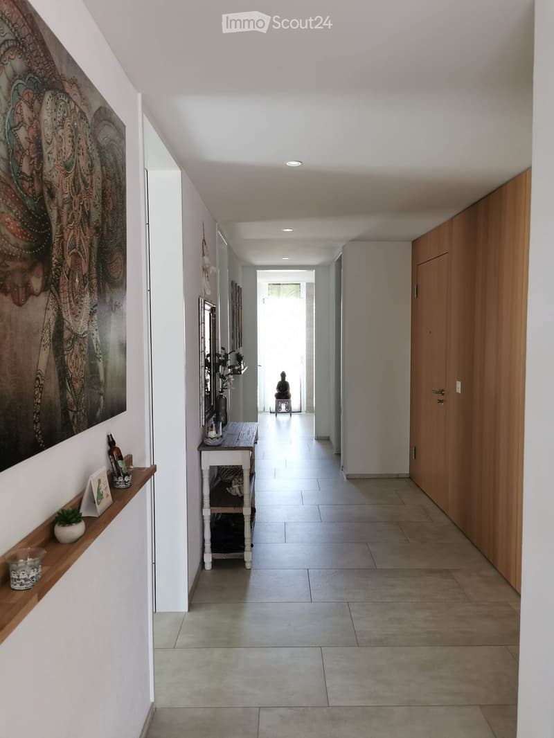 Modern hallway with white walls, tiled floor, wooden shelves, and artwork. Features a small table with decorative items and a small plant.