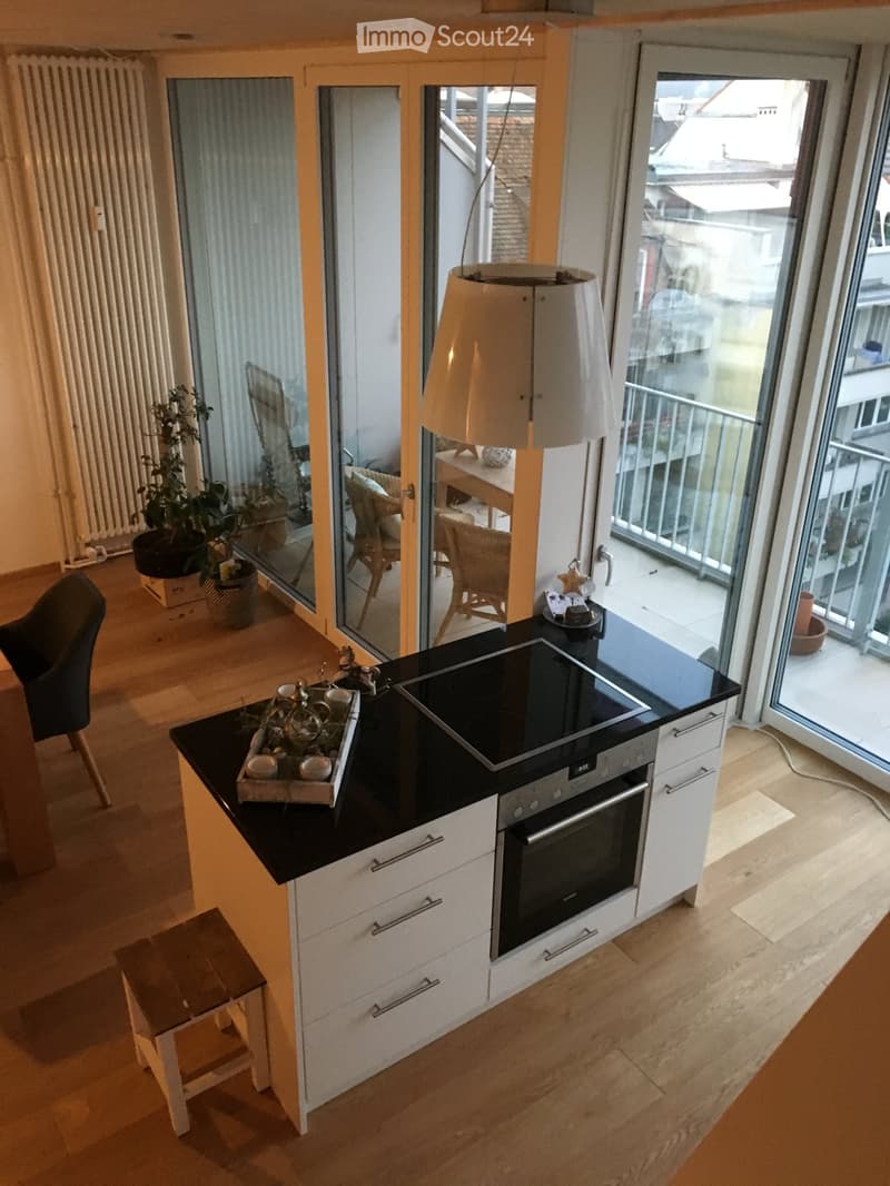 Modern kitchen with white cabinets, black countertops, built-in oven, and island. Features a hanging lamp, small table with chairs, and potted plants near the balcony.
