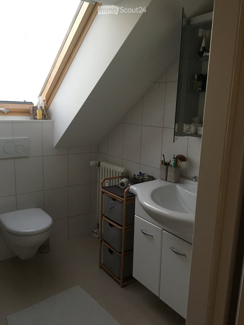 Modern attic bathroom with grey tiles, a toilet, a sink with a mirror, a radiator, and a small wooden storage unit.