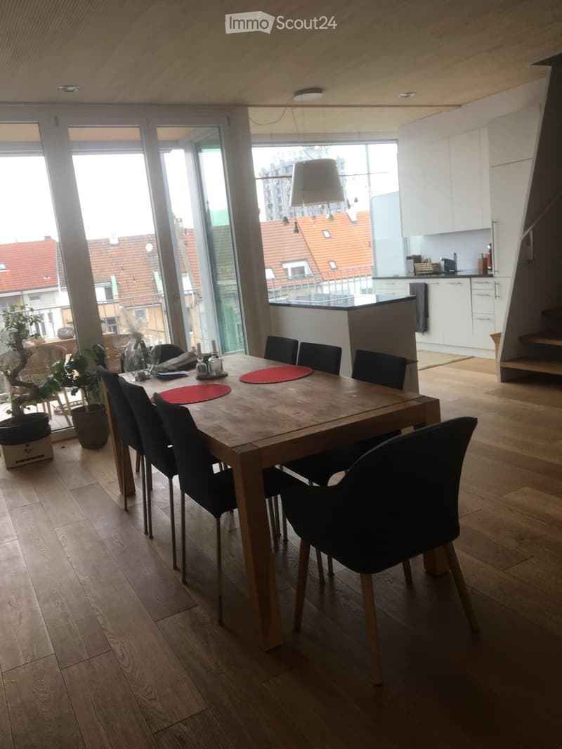Bright dining area with a wooden dining table, six black chairs, and a view of the kitchen area with white cabinets and a staircase leading upwards.