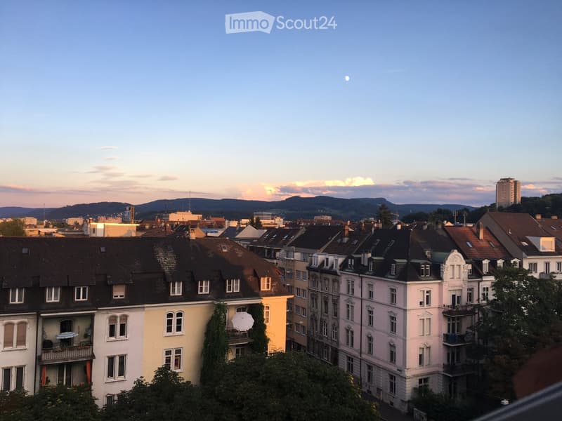 A cityscape view at dusk with multiple residential buildings, balconies, and a distant mountain range under a clear sky.