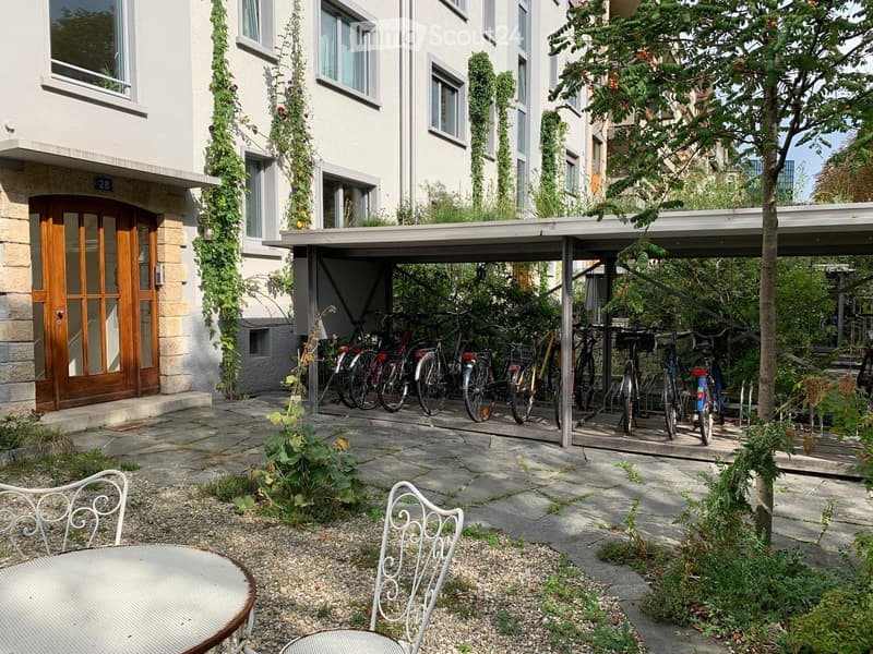Garden area with stone-paved ground, metal chairs, a round table, bicycles under a covered area, and a building with white walls and large windows.