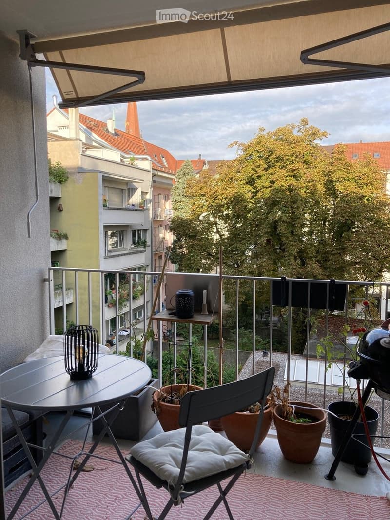 Balcony with a round table, chair, candle holder, and several potted plants, overlooking a cityscape with buildings and trees.