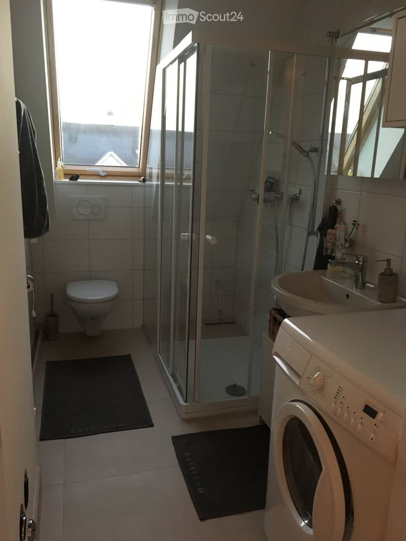 Modern bathroom with a toilet, sink, mirror, shower, and washing machine. Features tiled walls, a window, and two black bathroom rugs.