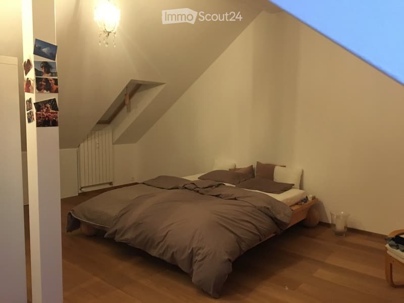 Cozy attic bedroom with wooden flooring, slanted ceilings, and a large bed with dark bedding. Includes a nightstand, small table, radiator, and picture frames on the wall.
