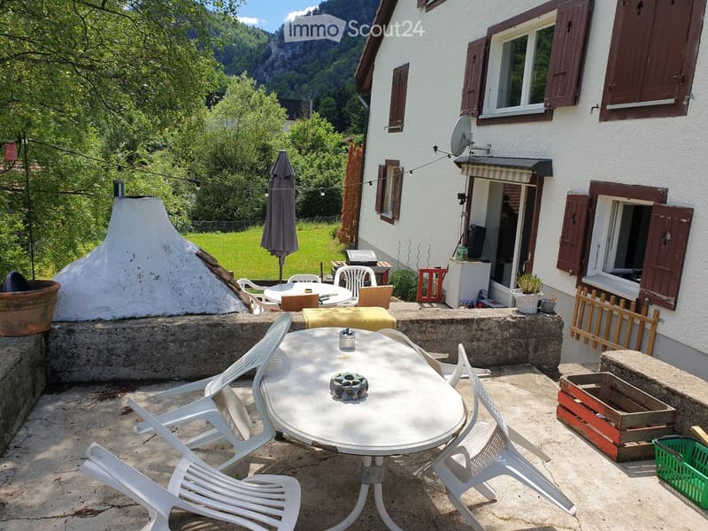 Outdoor patio area with a round table, white chairs, umbrella, potted plants, and a view of a green landscape and mountains.