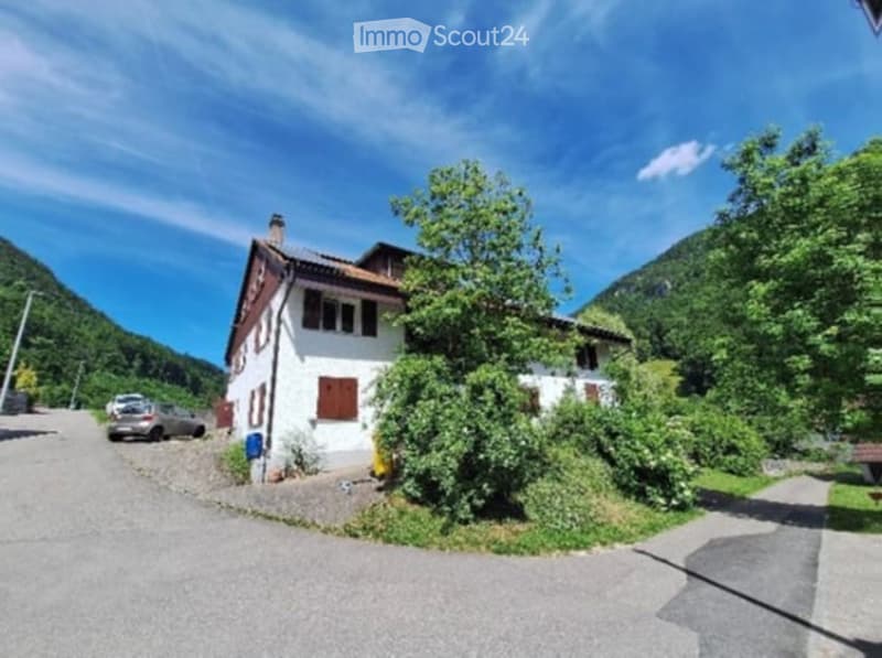 A two-story white house with brown shutters and a tiled roof, situated in a scenic, green valley with mountains in the background. The house has a small driveway with a parked car and is surrounded by lush vegetation.
