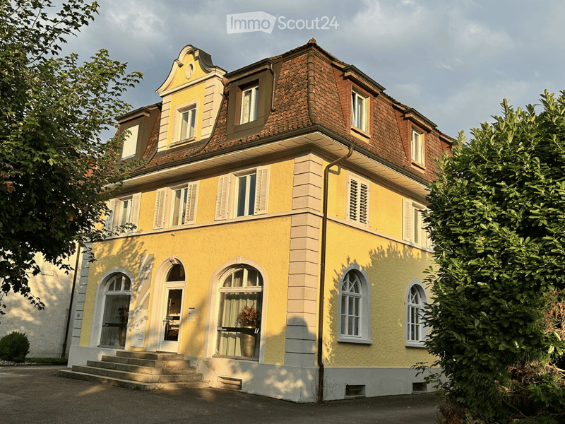 Three-story house, yellow walls, brown roof, white windows with shutters, arched windows, front door, bushes, steps leading to the entrance