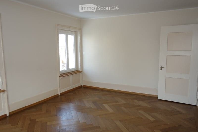 empty room with wooden parquet flooring, white walls, large window with wooden sill, closed door, and door frame
