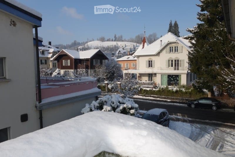 House covered in snow, white exterior, balcony, two cars on street, one car parked in front